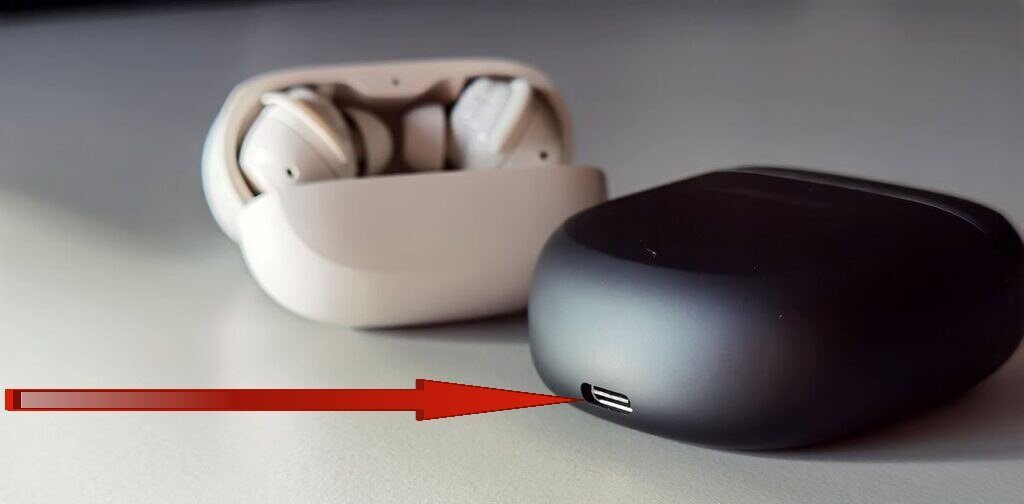 A red arrow pointing towards the charging port of one of the two earbuds present on a white surface