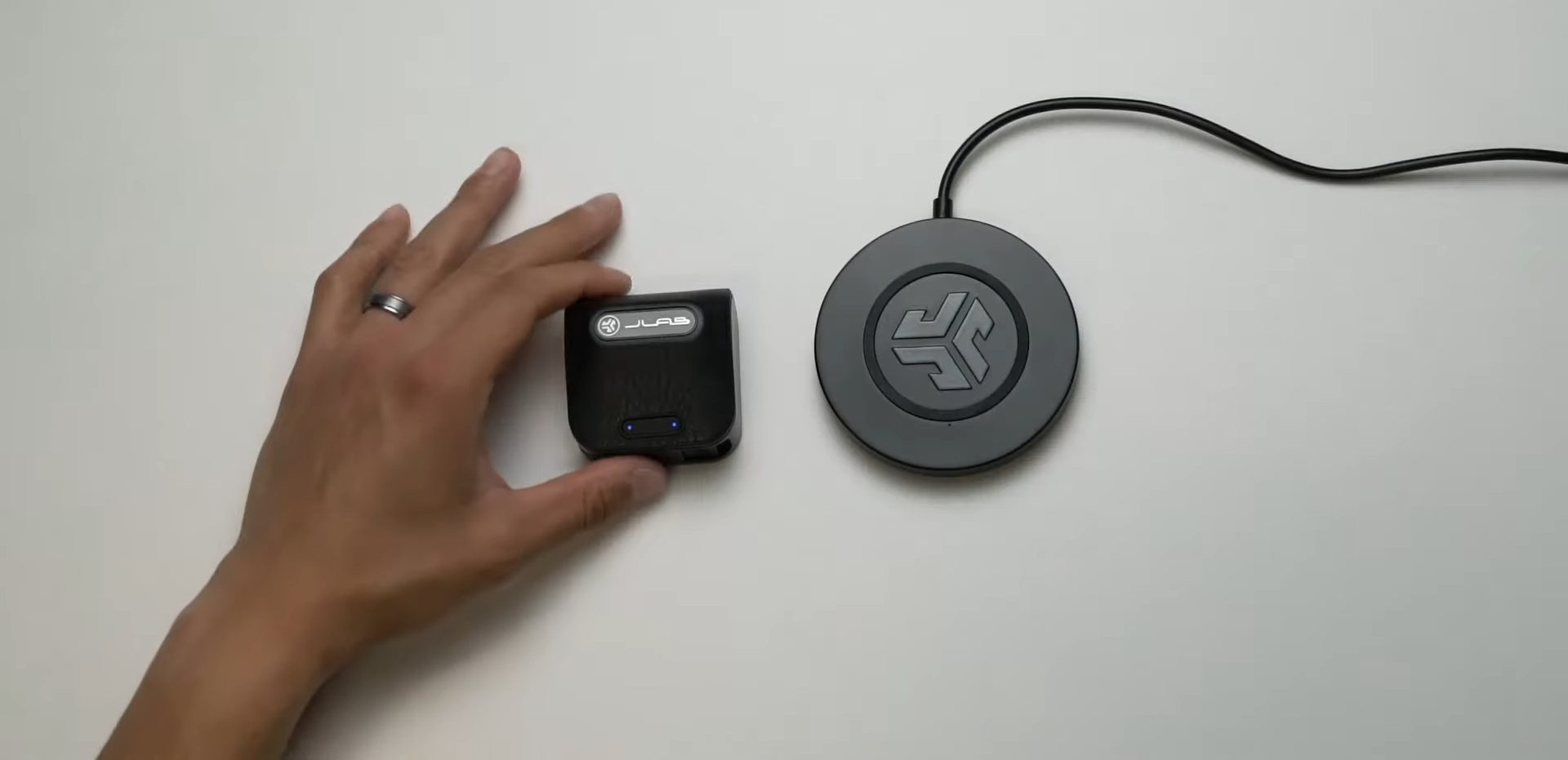 A person holding Jlab earbuds placed on a flate grey surfece while wireless charger is placed along side